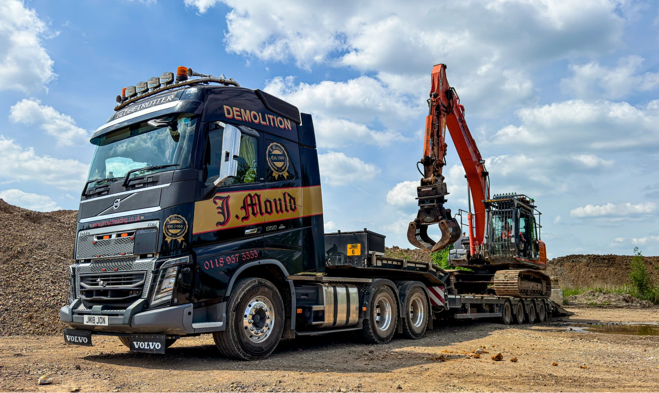 Black J Mould Volvo low loader carrying an orange excavator with a sorting grab, parked by aggregate stockpiles