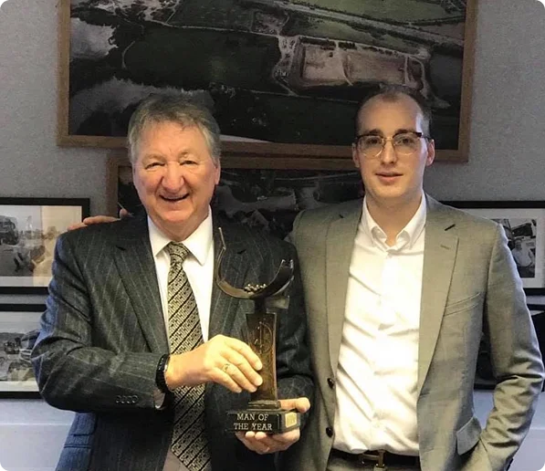 Two men in suits, the older one holding a 'Man of the Year' award, in front of framed aerial photos