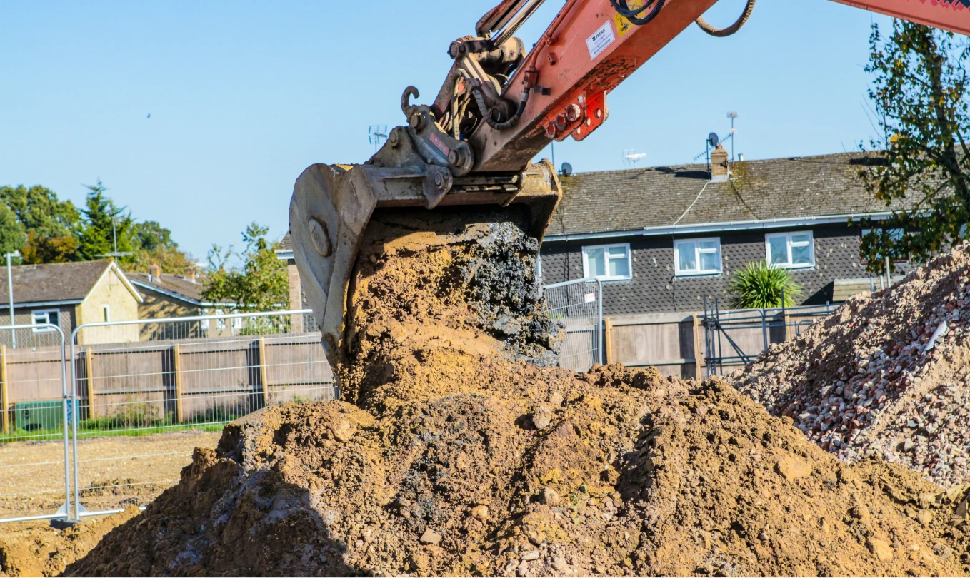 Orange J Mould excavator bucket tipping soil onto a heap beside crushed brick on a fenced site
