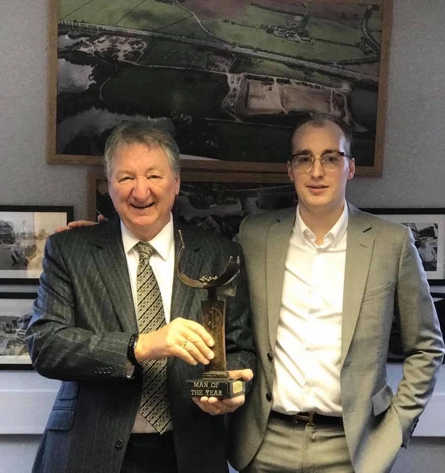 Two men holding a 'Man of the Year' trophy, aerial site photos on the wall behind