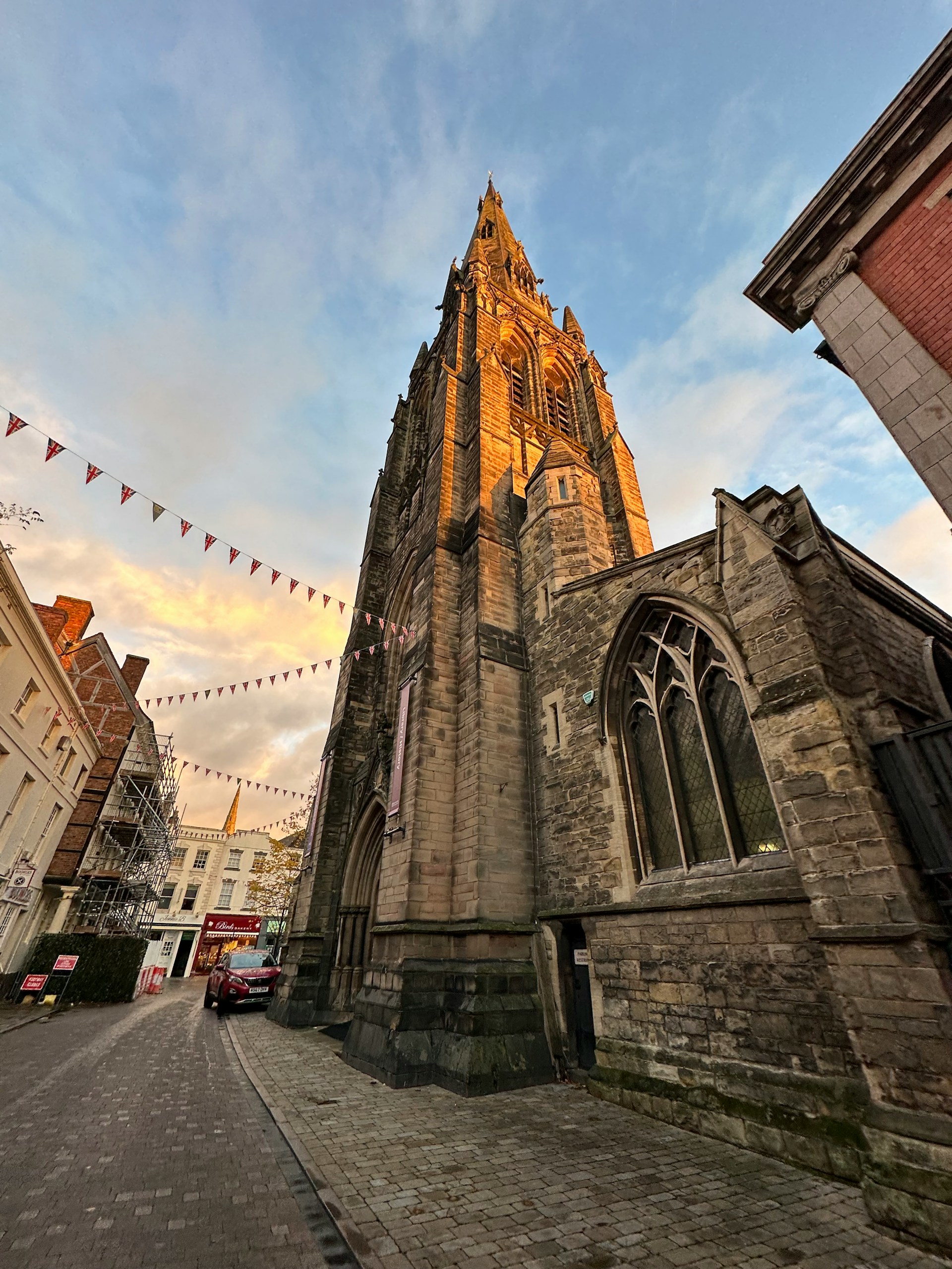 Sandstone church spire lit by the evening sun on a street hung with bunting, in an area J Mould covers for demolition