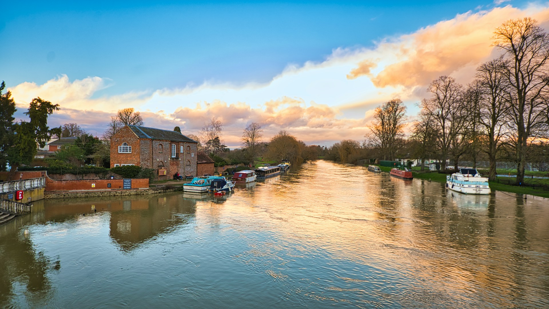 Boats moored on a wide river at sunset, with a brick riverside house and bare trees along the banks