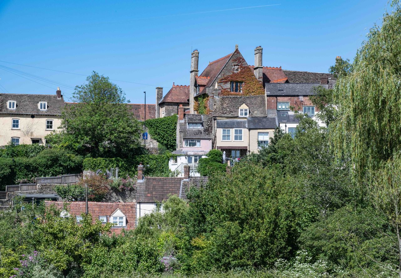 Stone and brick houses climbing a hillside above trees and a willow