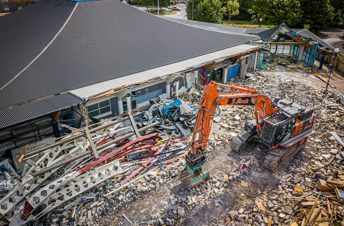 Aerial view of an orange J Mould excavator demolishing a large building with a curved metal roof