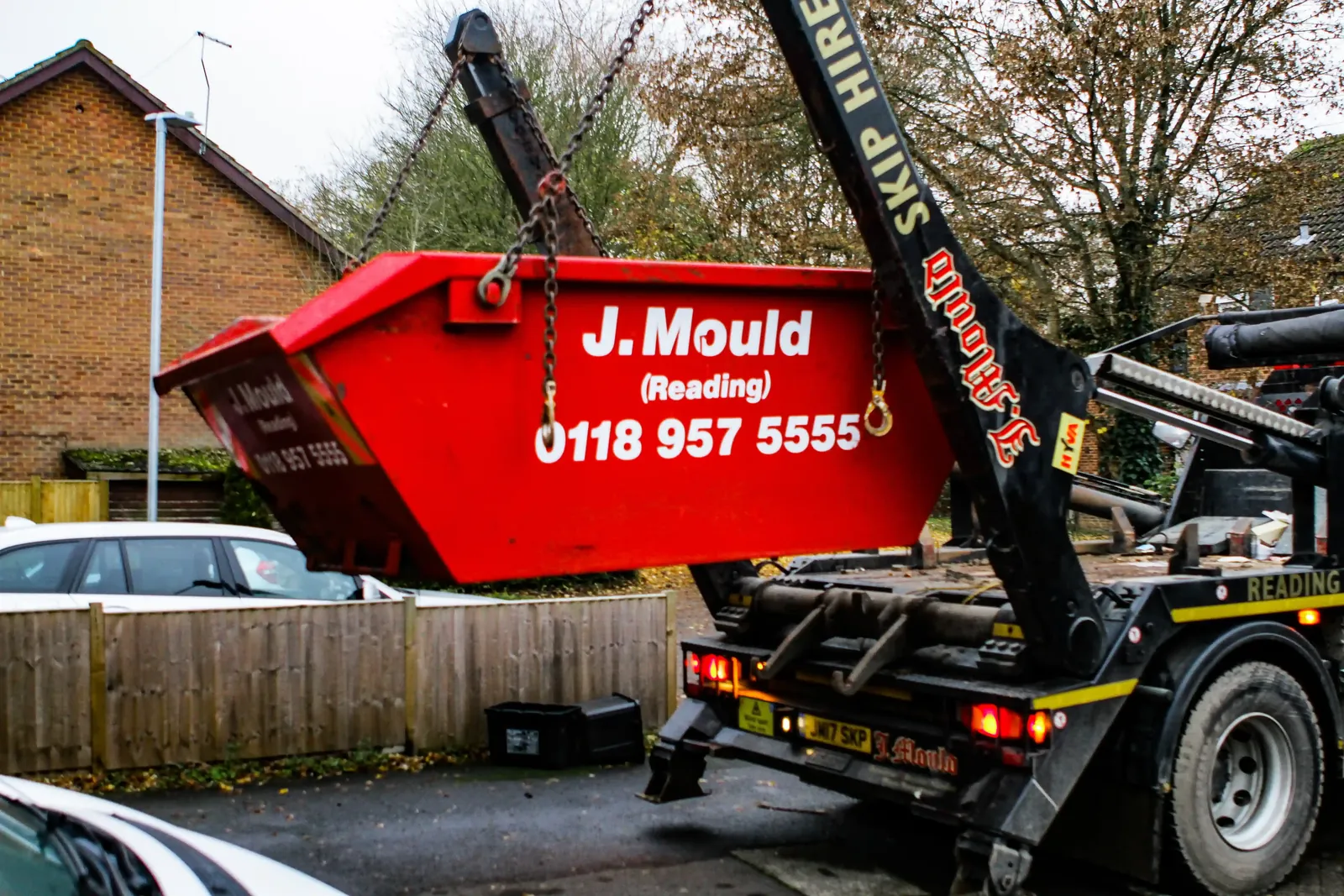 J Mould skip lorry lifting a red J Mould (Reading) skip on a residential street, part of J Mould's skip hire in Reading
